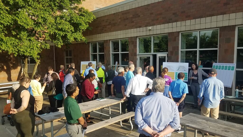 A crowd of community members gathered around picnic tables, trees, and display boards at a community input meeting for the Complete Streets policy