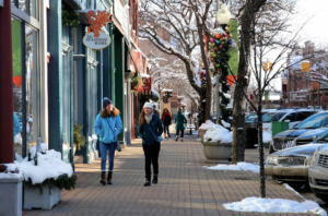 Two people wearing winter coats, scarves, and hats walk down a snowy sidewalk decorated for the holiday season.