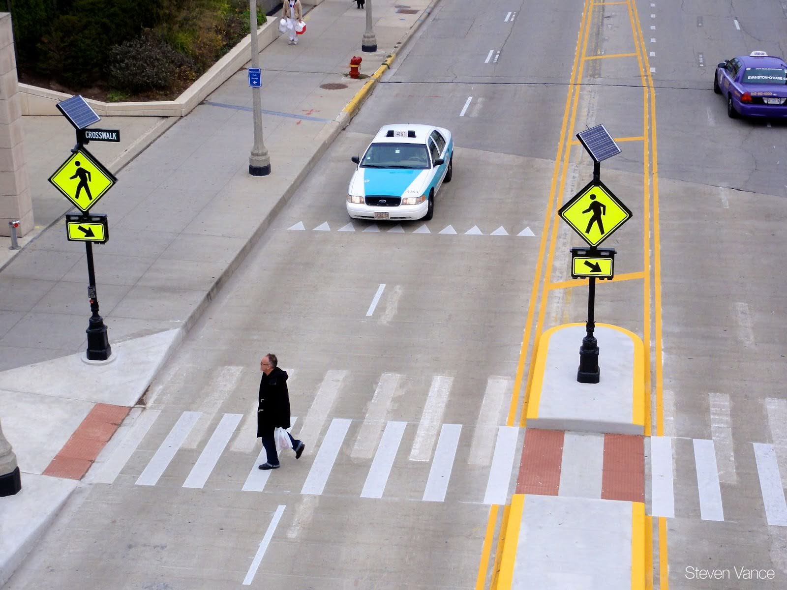 A pedestrian crosses a marked crossing that features two flashing rectangular beacons on either side of the street. A car slows to a stop several feet away.