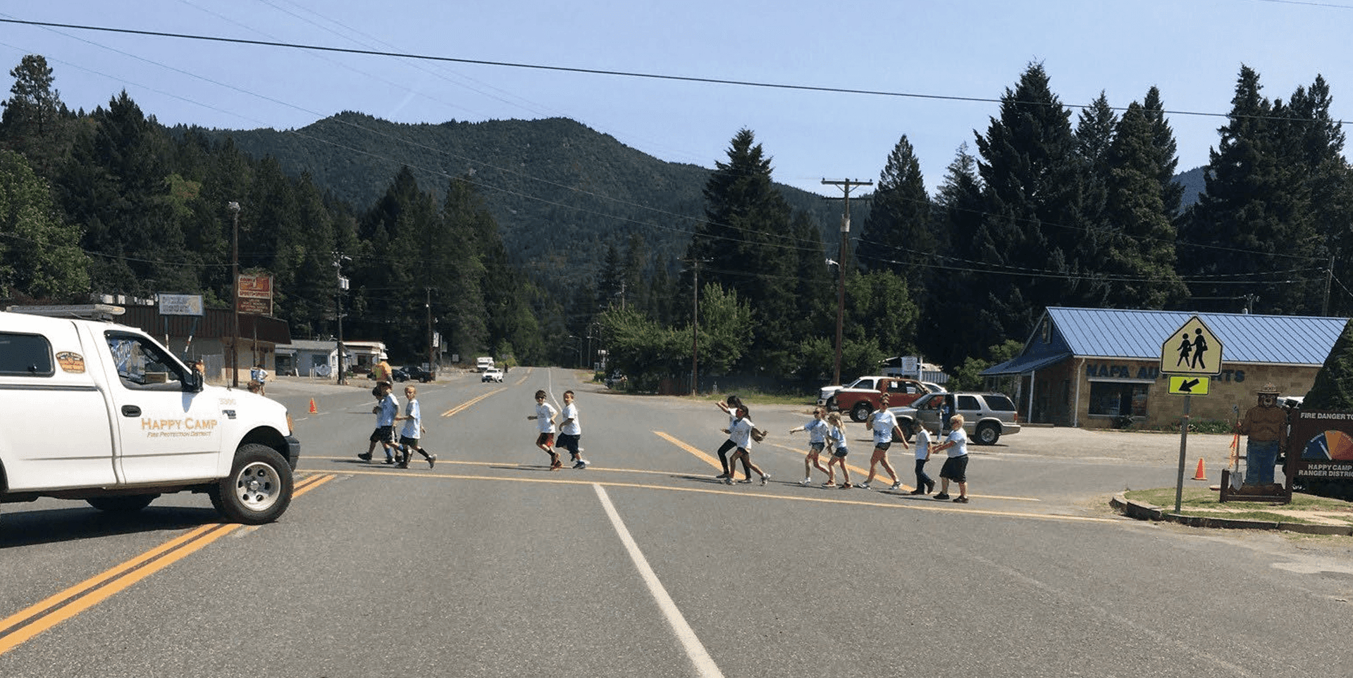 Happy Camp Complete Streets project before A group of children crosses a large intersection partially blocked by a white pick up truck with the logo "Happy Camp"