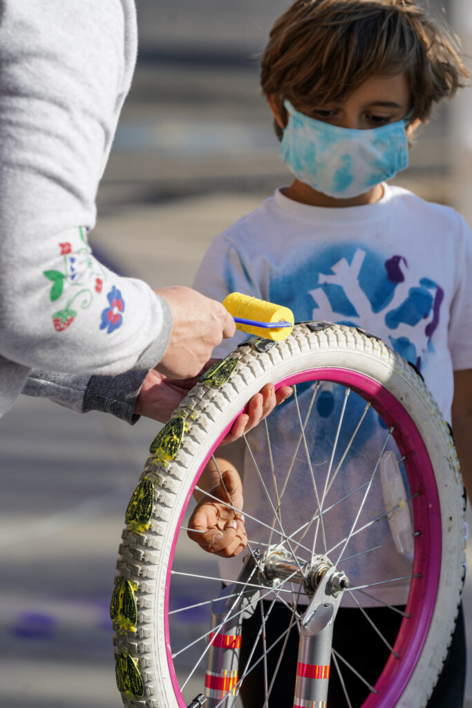Image of young person watching as an adult applies yellow paint to the footprints attached to the wheel of the play sculpture.
