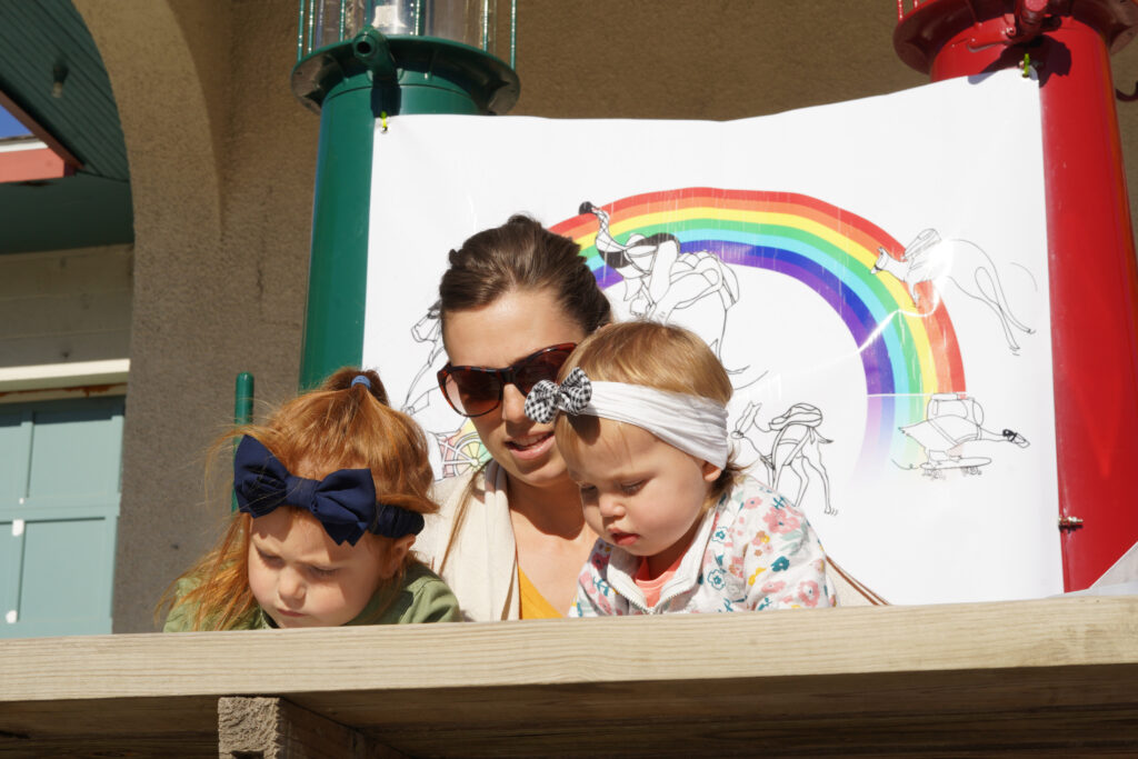 Image of two young people and an adult coloring papers. In the background is a poster featuring animals wearing masks.