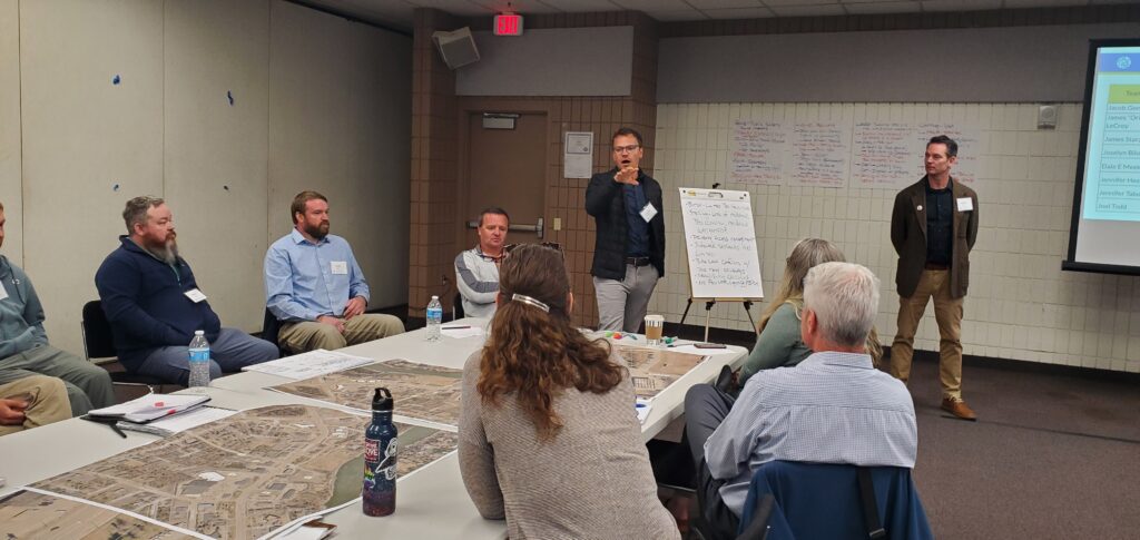 A man stands between an easel and a table full of maps of the intersection, as other people sit around the table, listening to what he has to say.
