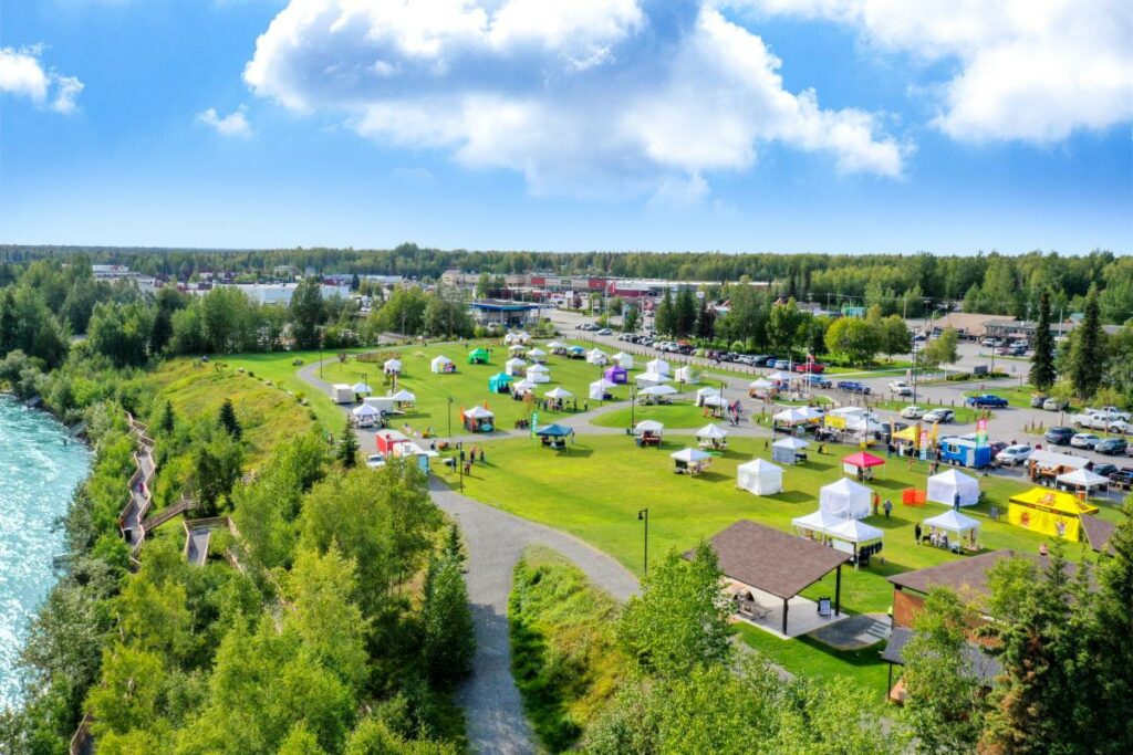 Rows of tents fill a grassy area lined with parked vehicles near the highway and a trail along the riverbank.