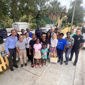 A group of people, representing a range of ethnicities, ages, and genders, stand and smile together on a West Palm Beach sidewalk