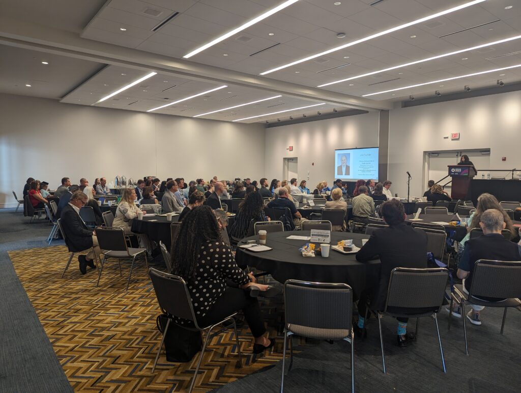 Conference Room with round tables and folks faceing a stage with a TB and Image of Tischler who is being honored with an award at the National Brownfields Training Conference. 