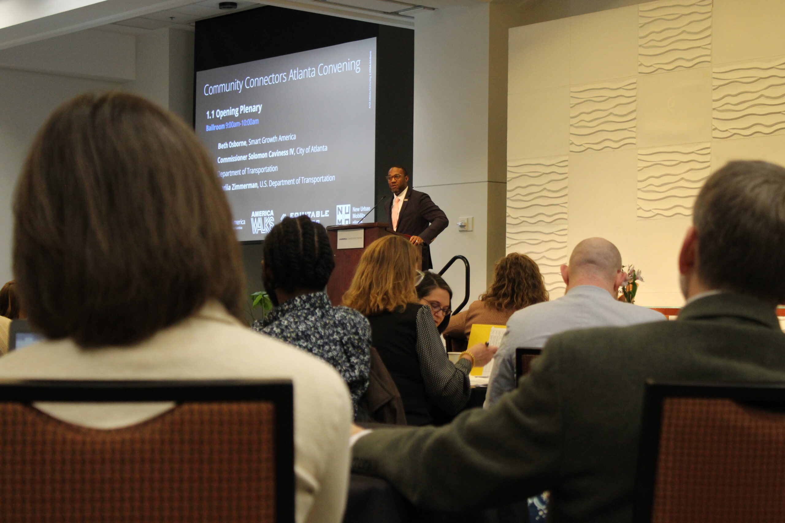 A speech is being given in front of a large crowd, in the background, a projector screen reads Community Connectors Atlanta Convening