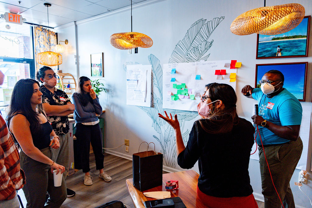 A diverse group of individuals, several wearing face masks, standing next to a wall with charts, engaged in a lively discussion with the group facilitator.