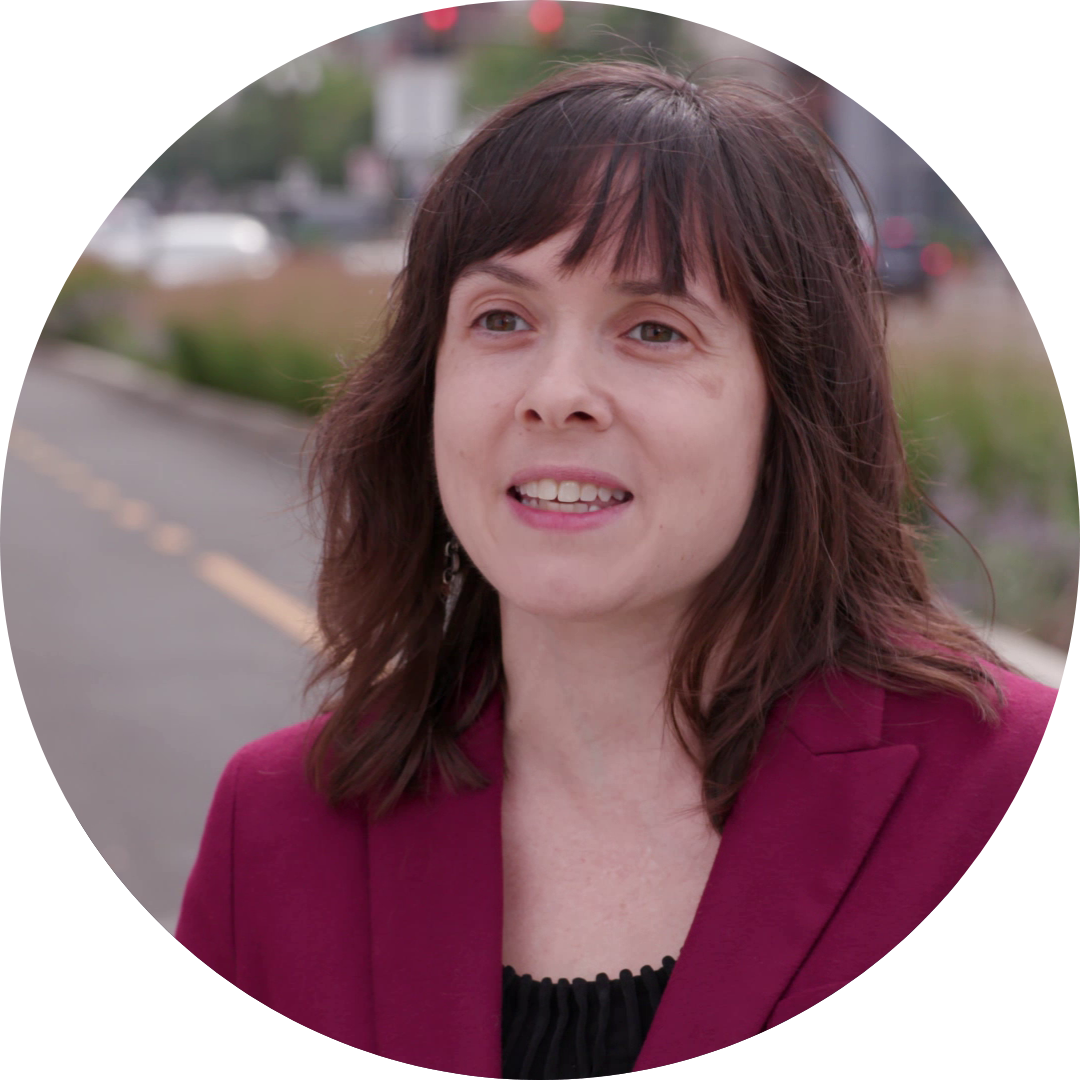 Jessica Cicchino's headshot, brown shoulder-length hair and bangs with a red blazer standing in front of a roadway.