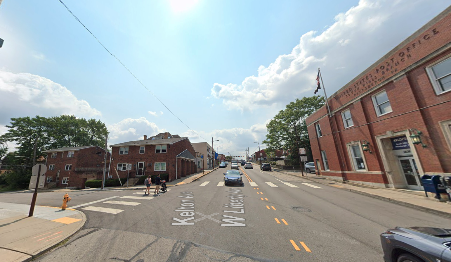 A young couple with a stroller crosses Kelton Avenue on the way to the West Liberty Avenue intersection, where a car is already passing by. To the right of the street is a post office. Other businesses rest on the left side of the street.