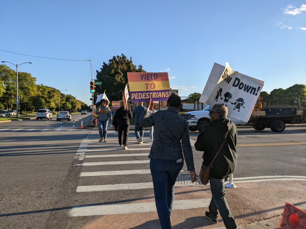 As they cross the street, people hold up signs calling for safer driver behavior, including a brightly-colored sign that says 