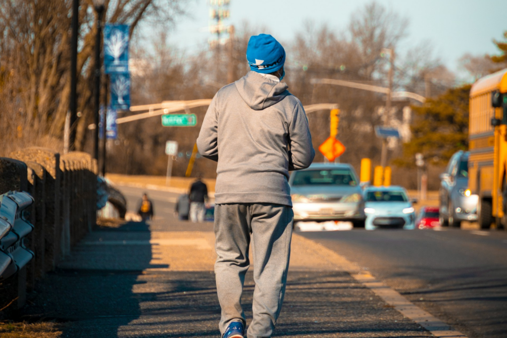 A Black man starts crossing a long intersection as rows of traffic on a wide road speed past him