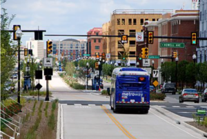 A blue DC metro bus travels down a two-land street with crosswalks. Yellow, orange, and red apartment buildings line theright side of the street.