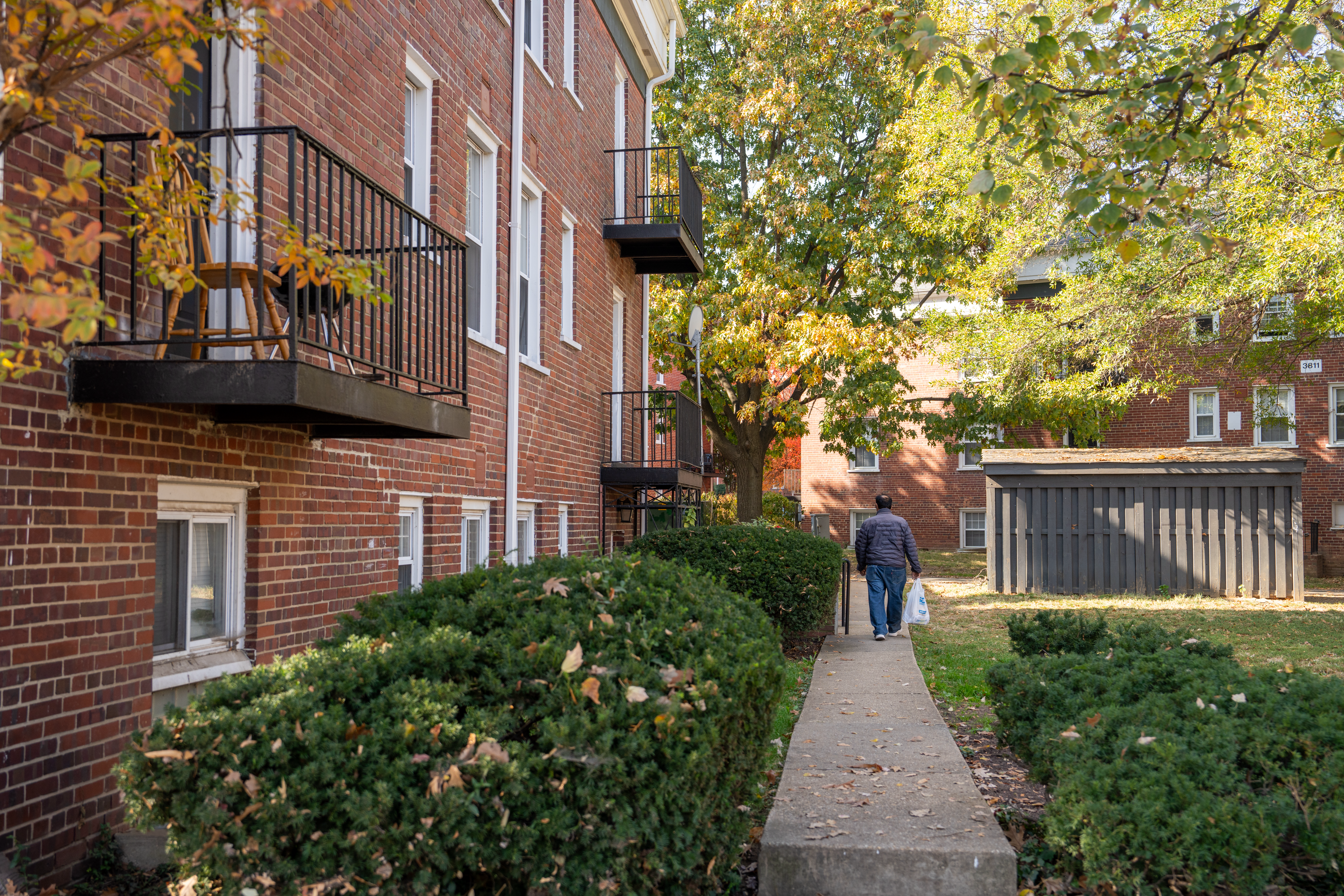 A man in a gray jacket walks down a shaded sidewalk surrounded by red brick apartment buildings
