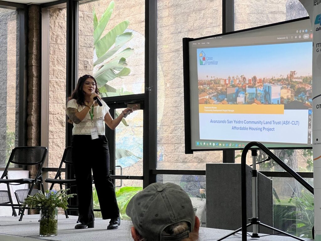 A woman in a white blouse and black slacks delivers a presentation from a sunlit stage