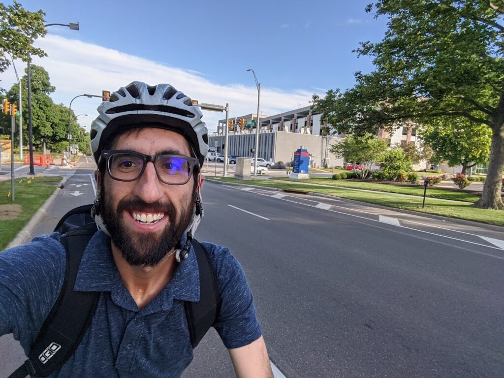 Man wearing a helmet appearing to ride a bike down a street lined with trees. 