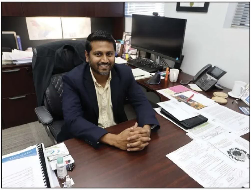 Man sitting and smiling up at the camera while sitting at a desk in an office. 