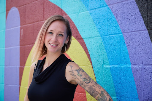 Woman smiling at the camera, standing in front of a colorful mural on a brick wall.