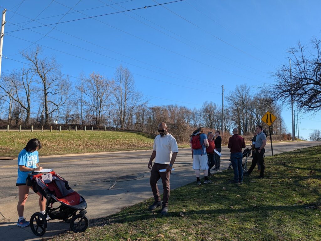 A group of eight people walks along Lakeshore Drive during a walk audit. The roadway has grass shoulders and no visible crosswalks.
