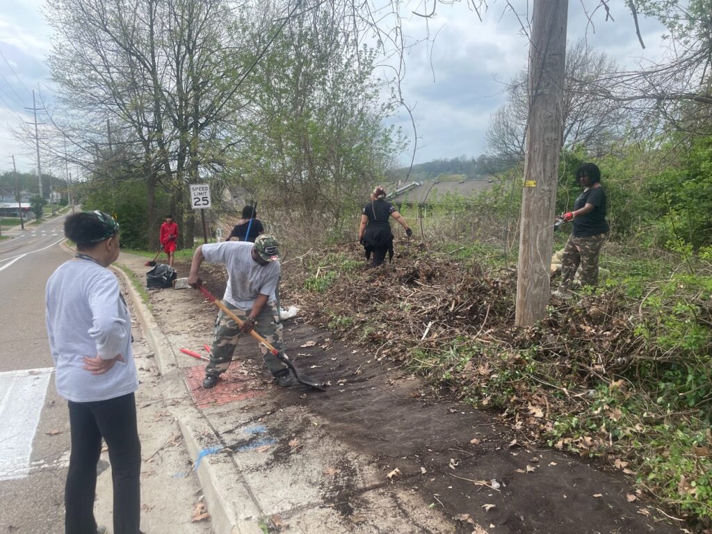 Six people using shovels and other tools to clear brush from a sidewalk beside a road. Part of the walkway is already cleared to bare dirt.