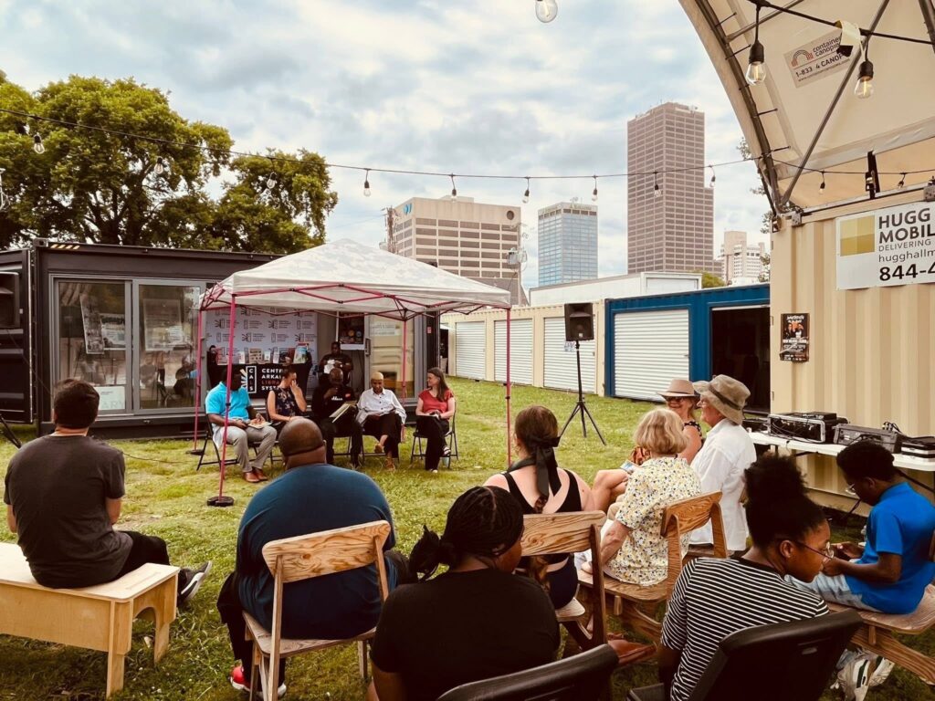 A crowd seated in folding chairs listens to a five-person panel speaking under a tent at an outdoor event.