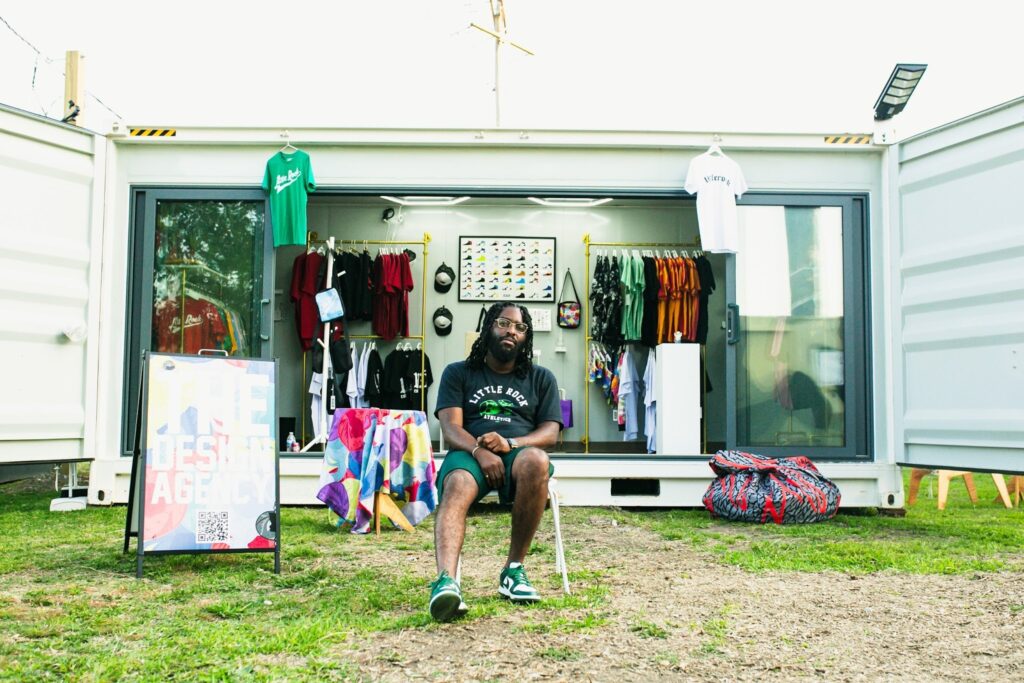 A business owner sits in a folding chair on the grass in front of their shipping container storefront at a pop-up market on West Ninth Street.